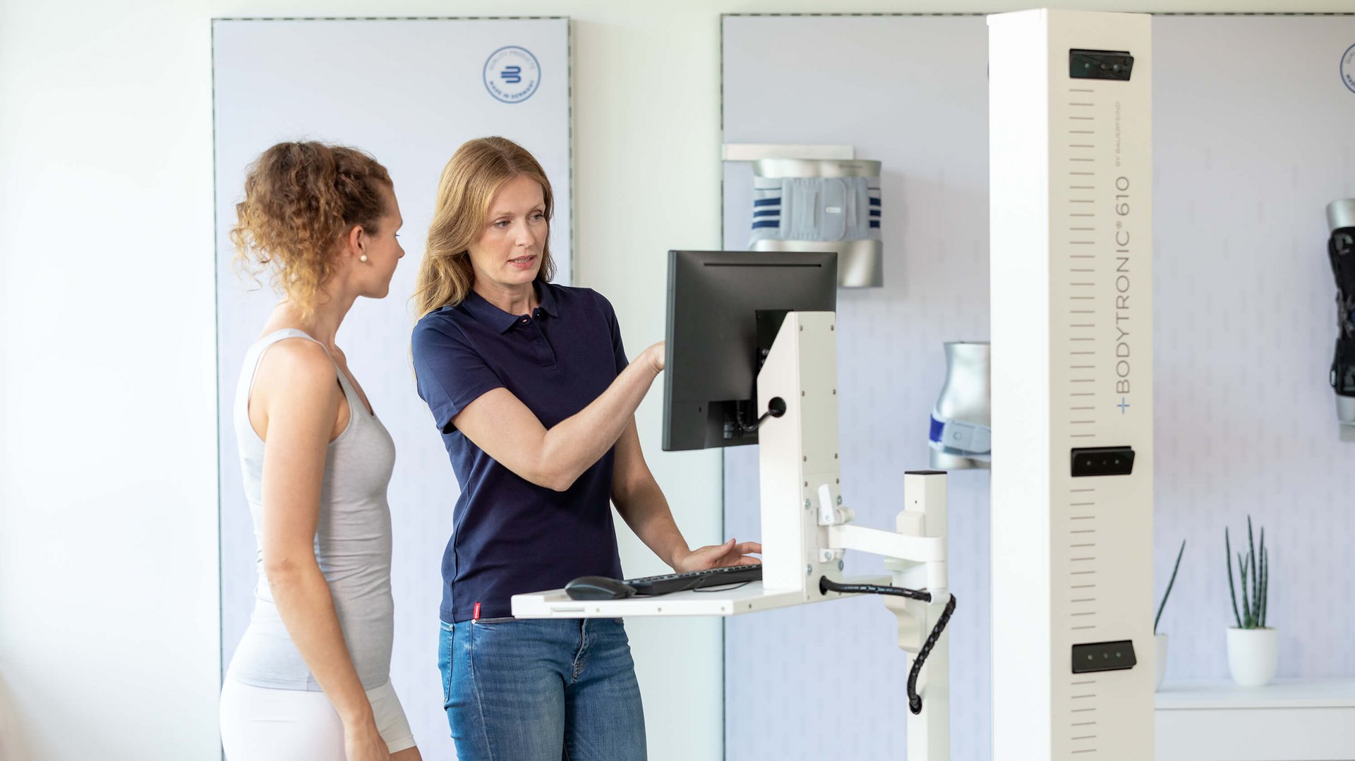 Zwei Frauen stehen neben einem Bodytronic-610-Messgerät in einem modernen Untersuchungsraum. Die eine Frau in Sportbekleidung schaut auf einen Bildschirm, während die andere, in Jeans und dunkelblauem Poloshirt, ihr etwas am Monitor erklärt.