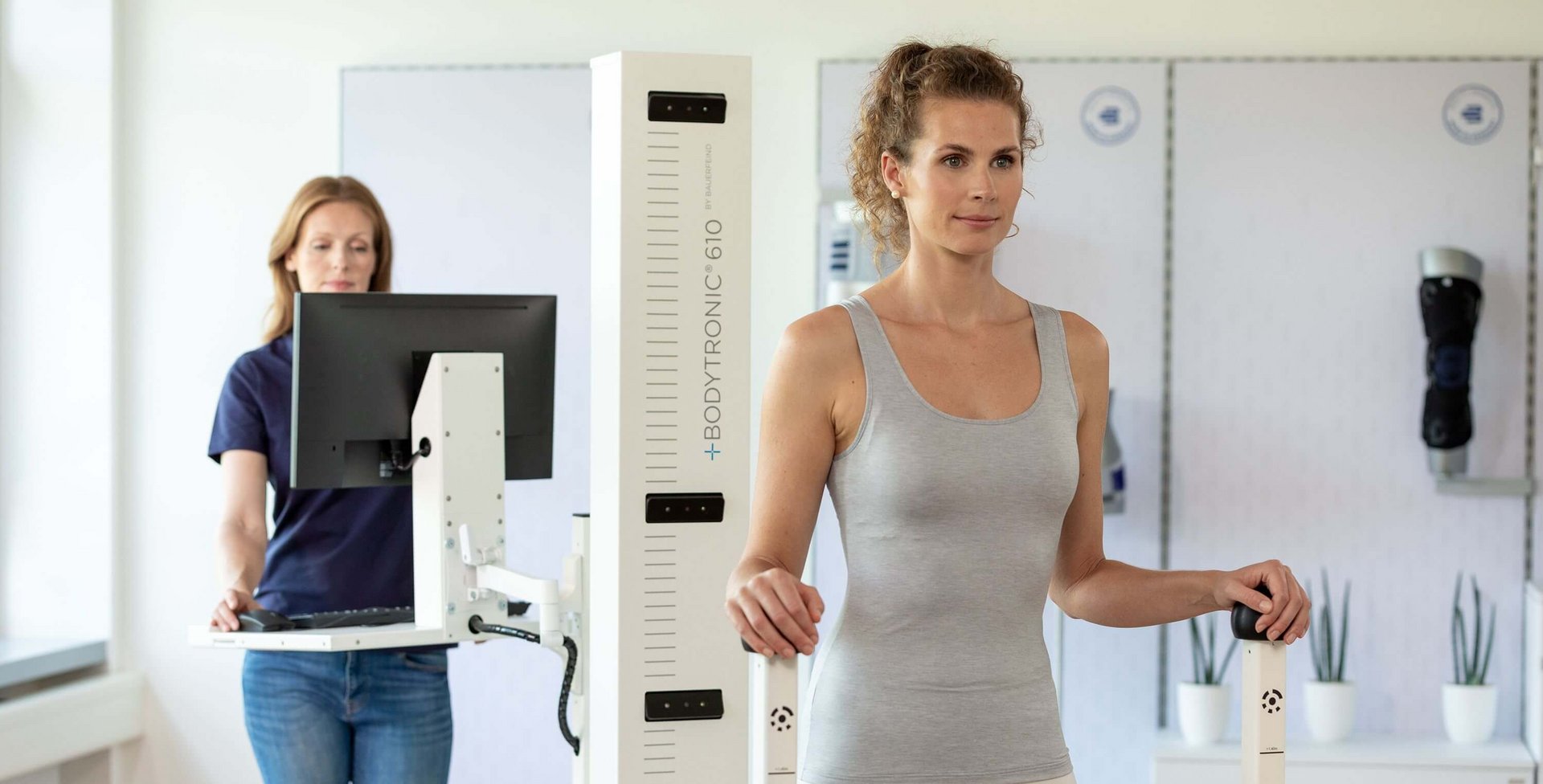 Frau in Sportbekleidung steht aufrecht an einem Bodytronic-610-Messgerät, das zur digitalen Körpervermessung dient. Im Hintergrund bedient eine Mitarbeiterin in Jeans und dunkelblauem Shirt einen Computer.